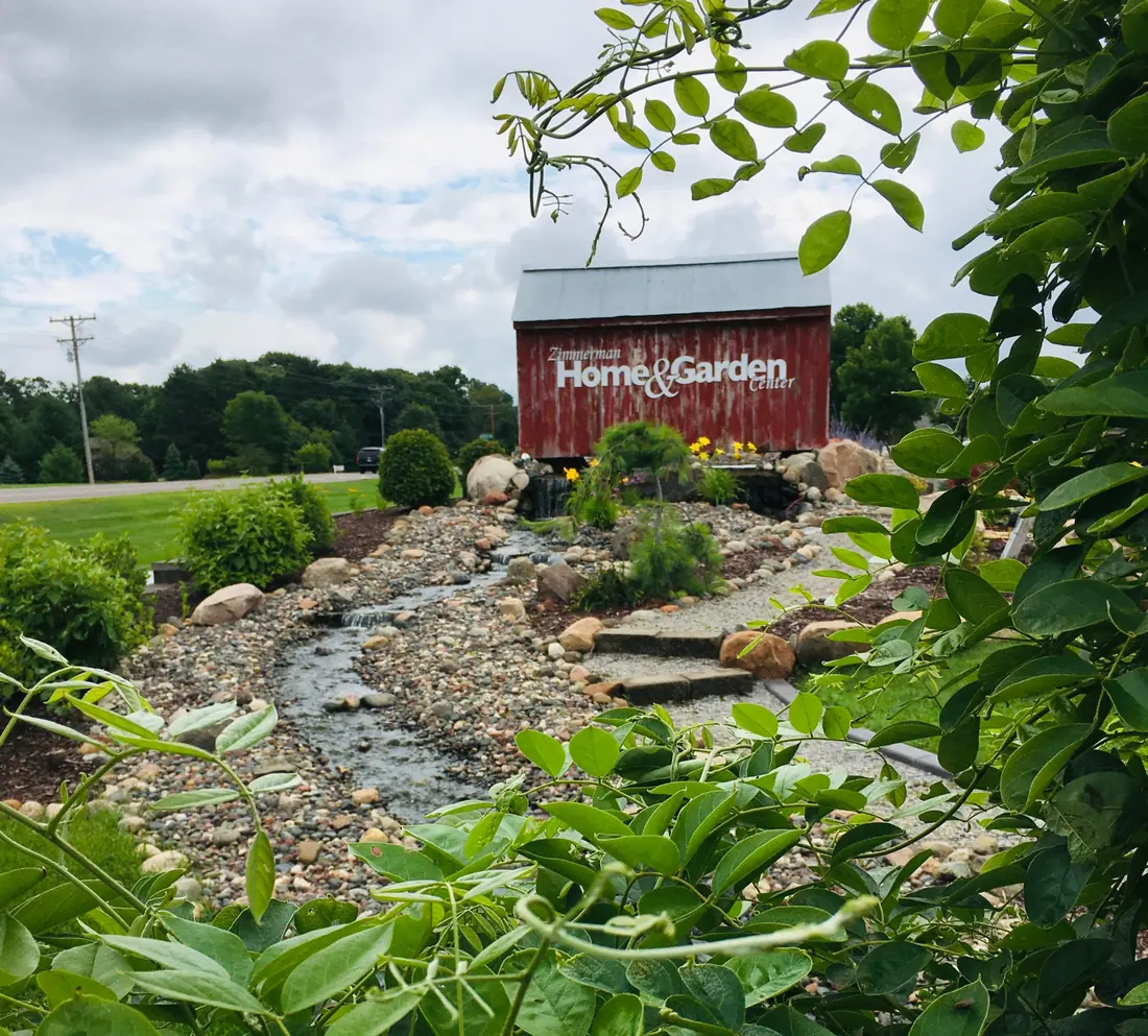 Red barn sign reading Home and Garden beside a stone-lined creek bed and plantings