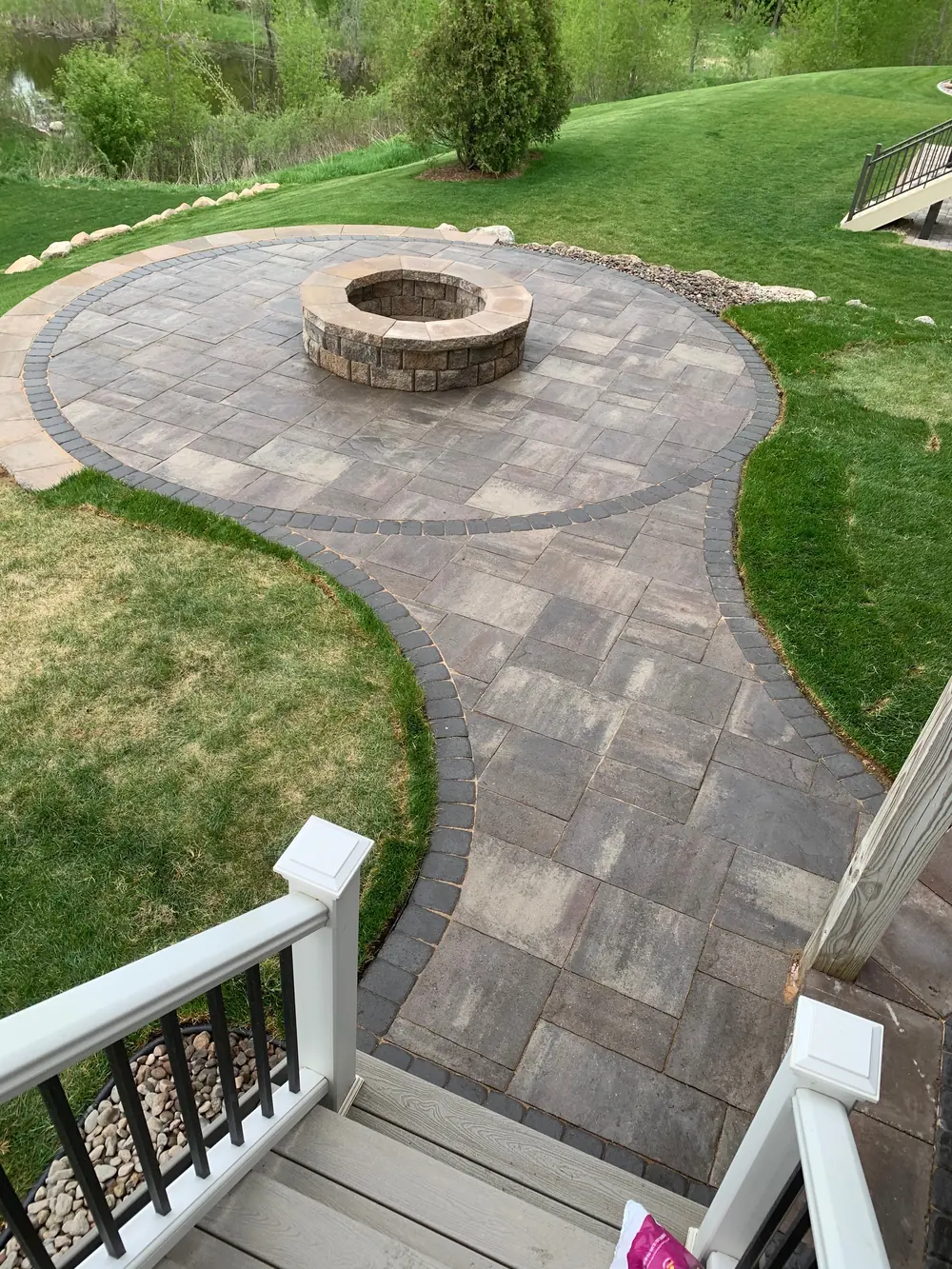 Aerial view of a paver patio with an in-ground fire pit beside a lawn and white railing