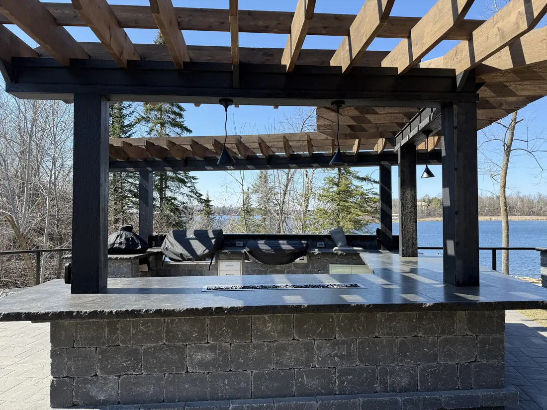 Dark timber pergola over a stone outdoor kitchen on a deck overlooking a lake